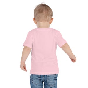 May include: A light pink toddler t-shirt with short sleeves. The shirt is made of a soft material and is paired with blue denim jeans. The child is viewed from the back, with their arms slightly outstretched.