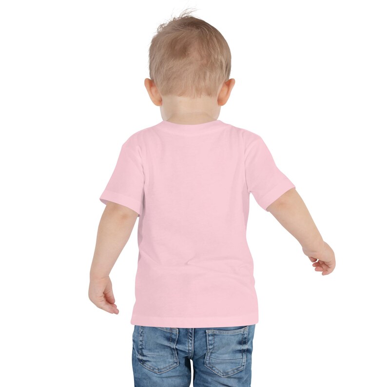 May include: A light pink toddler t-shirt with short sleeves. The shirt is made of a soft material and is paired with blue denim jeans. The child is viewed from the back, with their arms slightly outstretched.
