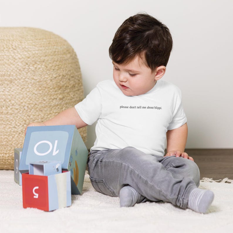 May include: A white short-sleeve t-shirt with the text "please don't tell me about blippi." A young child is wearing the shirt and sitting on a white rug. The child is playing with colorful building blocks.
