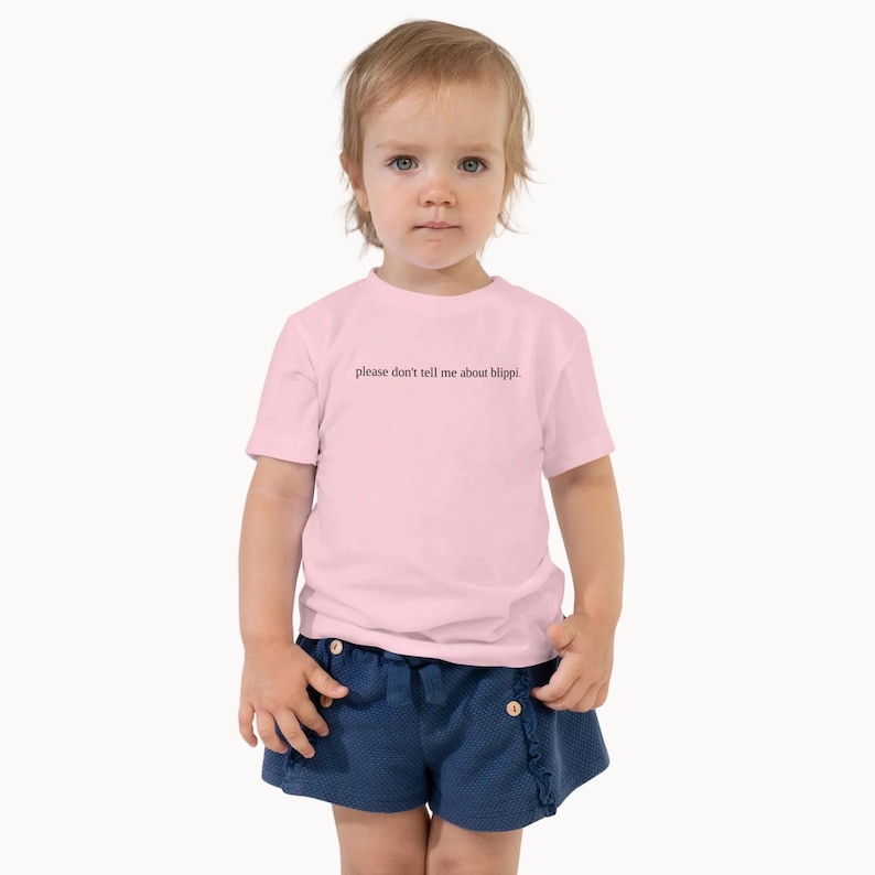 May include: A light pink toddler t-shirt with the text "please don't tell me about blippi" in black. The child is also wearing blue shorts. The shirt is made of a soft material.