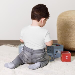 May include: A child wearing a white t-shirt, gray jeans, and striped socks sits on a white rug. The child is playing with a set of colorful wooden blocks. A woven ottoman is in the background.