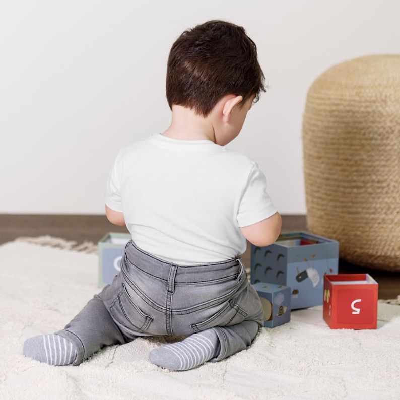 May include: A child wearing a white t-shirt, gray jeans, and striped socks sits on a white rug. The child is playing with a set of colorful wooden blocks. A woven ottoman is in the background.