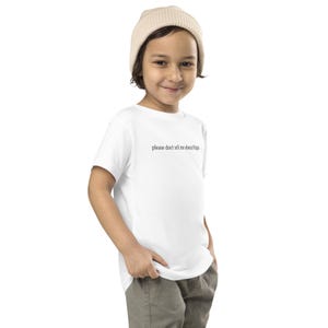 May include: A white t-shirt with the text "please don't tell me about blippi" in a black, simple font. The shirt is worn by a child, who is also wearing a beige beanie and khaki pants.