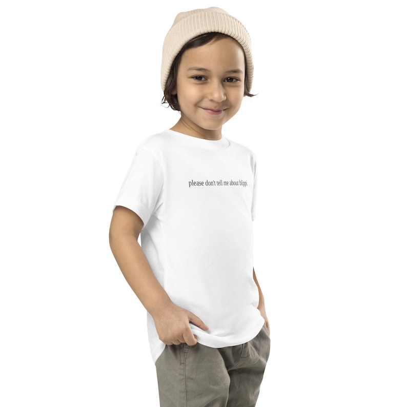 May include: A white t-shirt with the text "please don't tell me about blippi" in a black, simple font. The shirt is worn by a child, who is also wearing a beige beanie and khaki pants.
