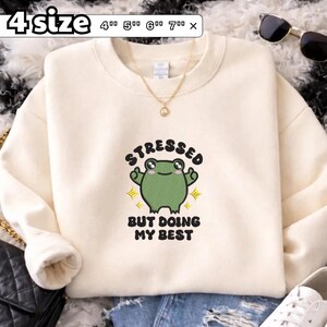 May include: A cream-coloured crewneck sweatshirt featuring an embroidered green frog graphic with the text "STRESSED BUT DOING MY BEST" and yellow star accents. The image includes a label indicating 4 sizes: 4", 5", 6", and 7" inches.