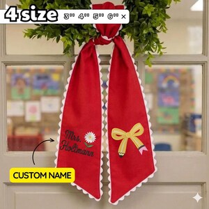 May include: A red fabric ribbon with white scalloped edges featuring the embroidered text Mrs. Holtmann, a white daisy, and a yellow pencil bow. The ribbon is available in 3 inch, 4 inch, 5 inch, and 6 inch sizes as indicated by the text.