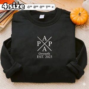 Może przedstawiać: Czarna bluza z okrągłym dekoltem i białym nadrukiem graficznym, na którym słowo PAPA ułożone jest w kształt krzyża. Poniżej grafiki znajduje się napis Gwyneth EST. 2023. Bluza dostępna jest w rozmiarach 4, 5, 6 i 7 cali.