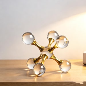 May include: A decorative jack-shaped sculpture featuring six clear glass spheres connected by a central gold-toned metal core. The object has a symmetrical, multi-directional design with smooth, reflective surfaces on both the glass and metal components.