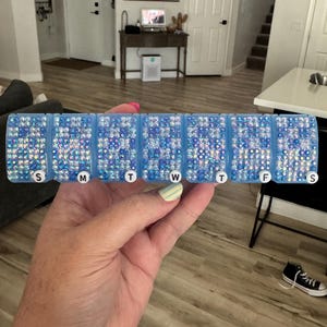 May include: A seven-compartment weekly pill organizer featuring a light blue plastic base. Each lid is covered in iridescent blue and clear rhinestones. The lids are labeled with white circular markers displaying the letters S, M, T, W, T, F, S.