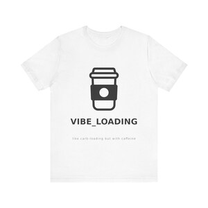 Könnte beinhalten: Weißes T-Shirt mit einem schwarzen Kaffeebecher-Motiv und dem Schriftzug "VIBE_LOADING" darunter. Der Text "like carb-loading but with caffeine" ist ebenfalls vorhanden. Das T-Shirt ist aus weichem Material.