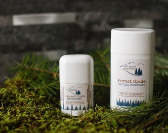 May include: Two white deodorant sticks with a forest glade scent. The smaller stick is 2.8 oz and the larger stick is 2.8 oz. Both sticks have a label with a mountain range and pine trees. The sticks are sitting on a bed of green moss.