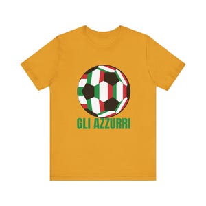 Puede incluir: Camiseta de color amarillo mostaza con un diseño de balón de fútbol con paneles verdes, blancos y rojos. El texto "GLI AZZURRI" está impreso en verde debajo. Una camiseta informal para los aficionados al fútbol.