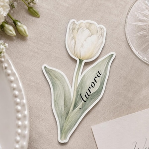 May include: A die-cut paper place card shaped like a white tulip with green leaves. The name Aurora is printed in black script along the stem and leaf. The illustration features soft watercolor textures and a white border outline.