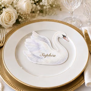 May include: A white swan-shaped place card featuring a watercolor illustration of a swan with soft grey and white feathers. The name Sophia is printed in elegant gold script across the center of the swan's body, resting on a white dinner plate.