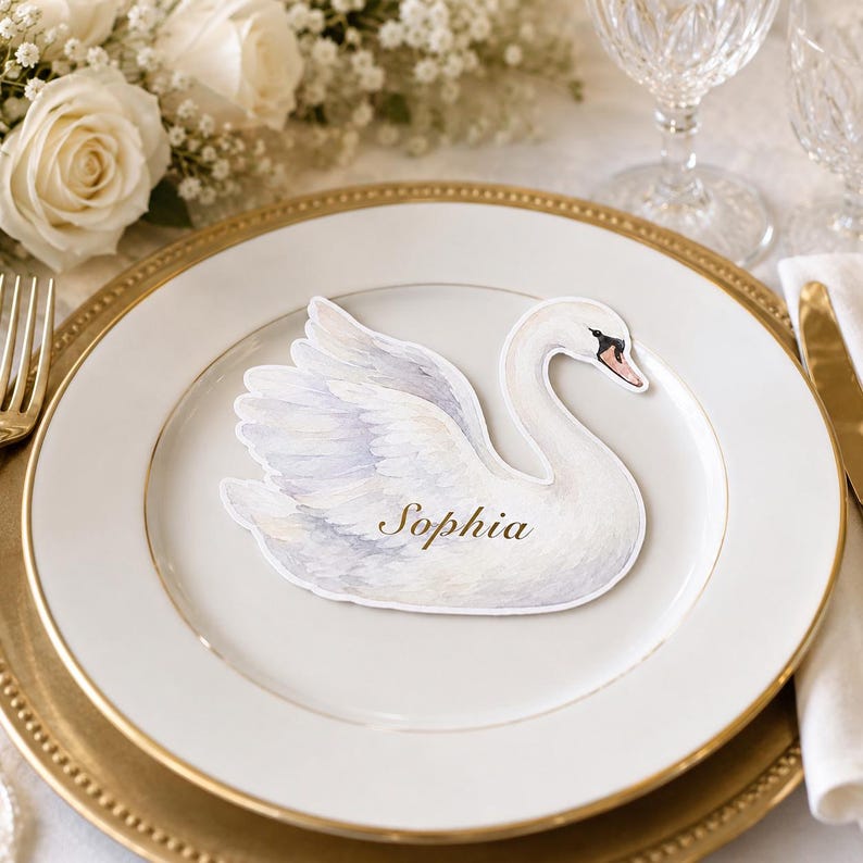 May include: A white swan-shaped place card featuring a watercolor illustration of a swan with soft grey and white feathers. The name Sophia is printed in elegant gold script across the center of the swan's body, resting on a white dinner plate.