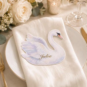 May include: A die-cut paper place card shaped like a swan with soft watercolor-style white and light purple feathers. The swan features a black and orange beak and the name Julia is printed in elegant gold script on the body of the swan.