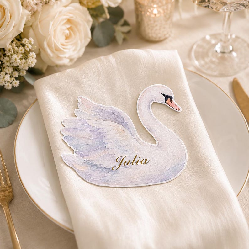 May include: A die-cut paper place card shaped like a swan with soft watercolor-style white and light purple feathers. The swan features a black and orange beak and the name Julia is printed in elegant gold script on the body of the swan.