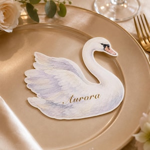May include: A die-cut paper place card shaped like a swan with watercolor-style feathers in shades of white, soft blue, and lavender. The name Aurora is printed in elegant gold script across the center of the swan's body on a gold plate.