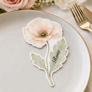 May include: A die-cut paper place card shaped like a pale peach poppy flower with a green stem and leaves. The name Rose is printed in black cursive script on one of the leaves. The flower features delicate petals and a central green stamen.