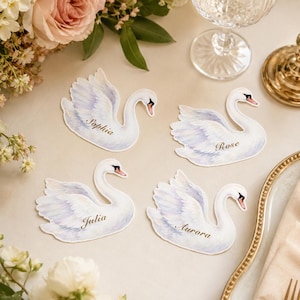 May include: Four die-cut paper place cards shaped like white swans with soft blue and purple shading on the wings. Each swan features a unique name printed in elegant gold script: Sophia, Rose, Julia, and Aurora, with black and orange beaks.