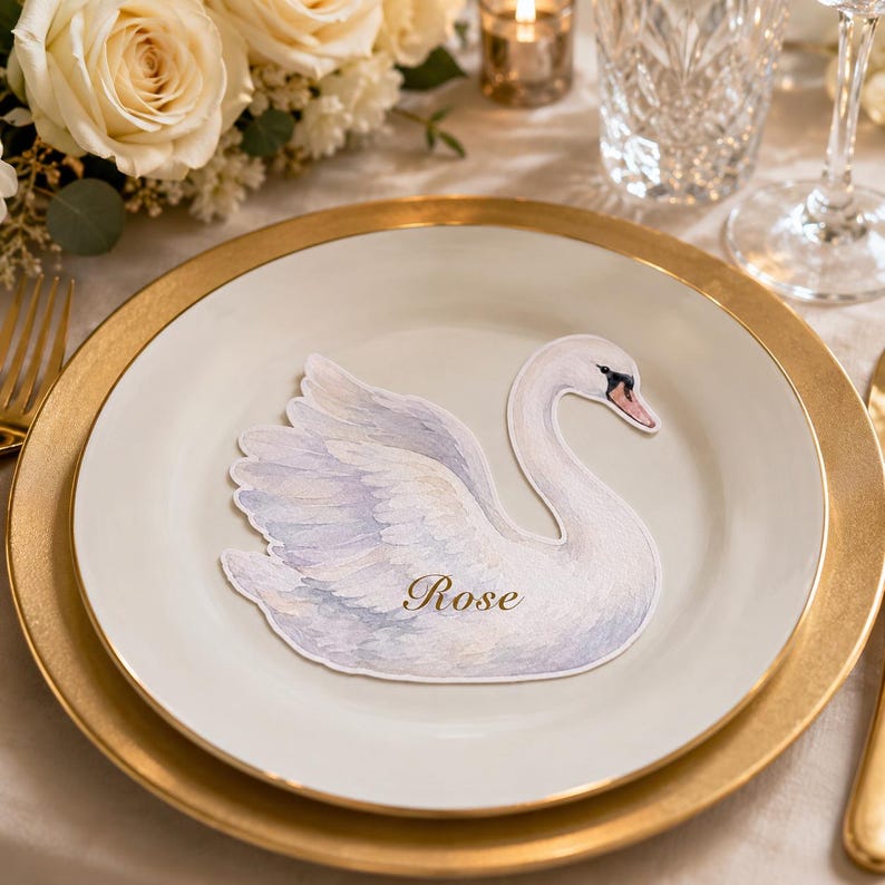 May include: A die-cut paper place card shaped like a swan with white and soft blue watercolor feathers. The swan features a black and orange beak and the name Rose is printed in elegant gold script across the center of the swan's body.