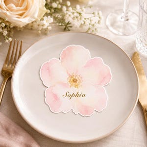 May include: A die-cut paper place card shaped like a five-petaled flower with a soft watercolor wash in shades of pale pink and white. The center features a yellow stamen detail, and the name Sophia is printed in a gold serif font on the petal.