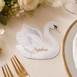 May include: A die-cut paper place card shaped like a swan with watercolor-style white and soft grey feathers, a black and orange beak, and the name Sophia printed in gold script lettering across the center of the swan's body.