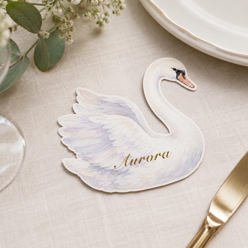May include: A die-cut paper place card shaped like a swan with watercolor-style white and soft purple feathers. The swan has a black and orange beak and the name Aurora is printed in gold cursive script across the center of the swan's body.