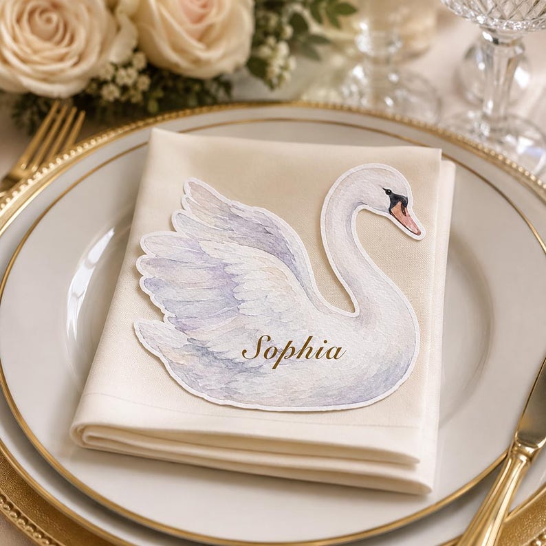 May include: A die-cut paper place card shaped like a white swan with soft grey and blue shading on the wings. The name Sophia is printed in a gold script font across the center of the swan body. The swan is resting on a folded cream-colored napkin.