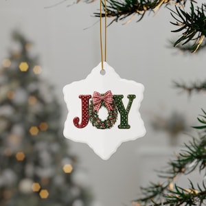 May include: A white snowflake-shaped ceramic ornament hanging from a gold string. The center features the word JOY in red and green with white polka dots. The letter O is replaced by a green wreath with a red and white gingham bow on top.