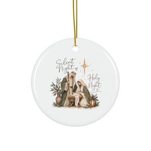 May include: A round white ceramic ornament with a gold hanging string. The center features a nativity scene illustration with a man and woman surrounding a baby in a manger, a lantern, and a vase. The text reads Silent Night Holy Night.