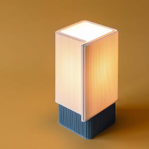 May include: A rectangular table lamp featuring a ribbed white shade that emits a warm glow, positioned atop a solid, dark blue, vertically ribbed base. The design consists of a clean, geometric structure with a distinct two-tone color palette.