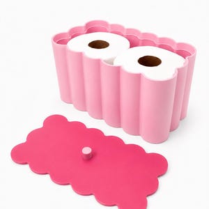 May include: A light pink rectangular toilet paper holder with a scalloped edge design, containing two rolls of white toilet paper. A separate, darker pink rectangular lid with a scalloped edge and a small central knob is placed in front.