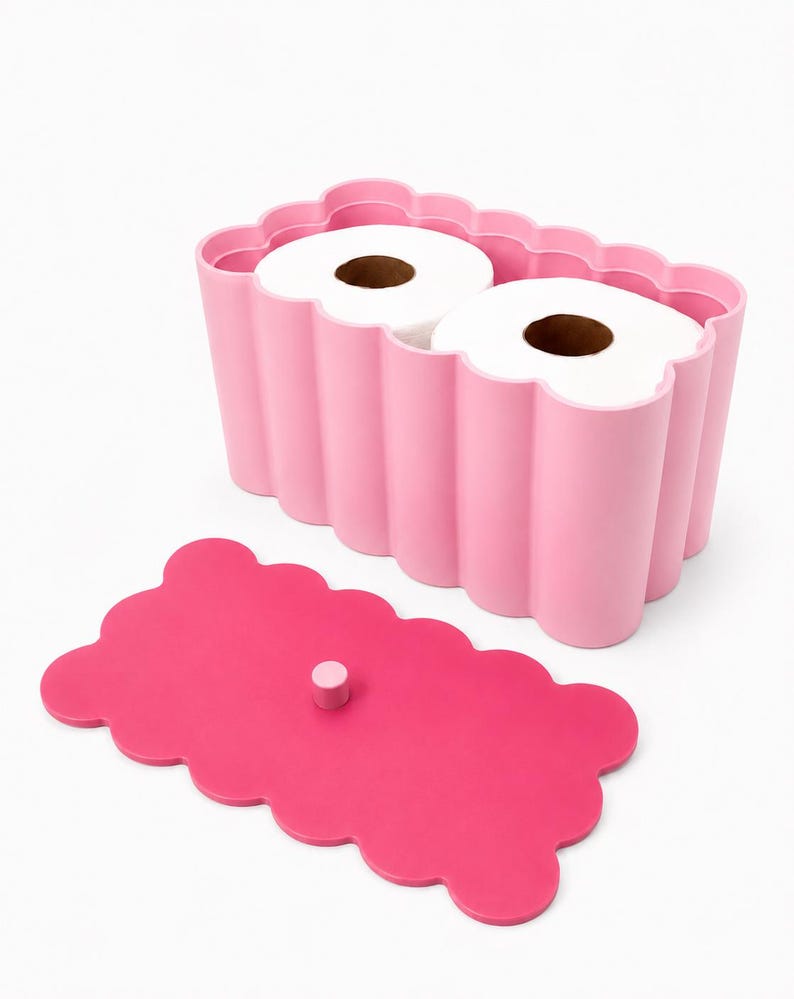 May include: A light pink rectangular toilet paper holder with a scalloped edge design, containing two rolls of white toilet paper. A separate, darker pink rectangular lid with a scalloped edge and a small central knob is placed in front.