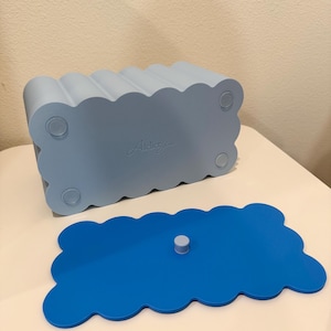 May include: A light blue rectangular storage box with a scalloped edge design and four circular feet on the base. The brand name Atelier Jun is embossed on the bottom. A separate, flat, bright blue scalloped lid with a small central knob is included.