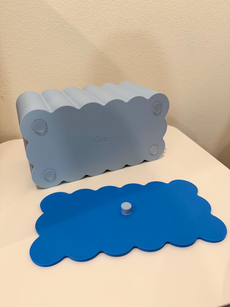 May include: A light blue rectangular storage box with a scalloped edge design and four circular feet on the base. The brand name Atelier Jun is embossed on the bottom. A separate, flat, bright blue scalloped lid with a small central knob is included.