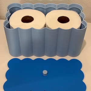 May include: A light blue rectangular storage container with a scalloped edge design holding two white toilet paper rolls. The container includes a separate, matching dark blue scalloped lid with a small central knob for easy removal and handling.