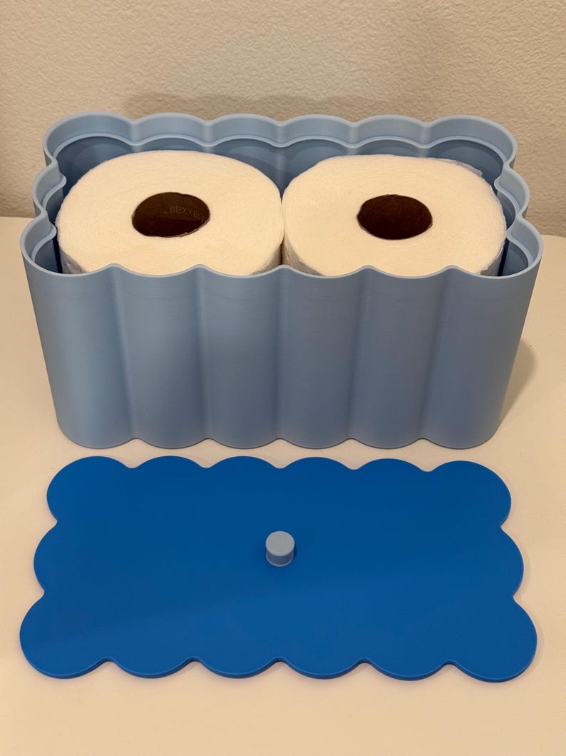 May include: A light blue rectangular storage container with a scalloped edge design holding two white toilet paper rolls. The container includes a separate, matching dark blue scalloped lid with a small central knob for easy removal and handling.
