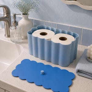 May include: A light blue rectangular toilet paper holder with a scalloped edge design, containing two rolls of white toilet paper. The holder includes a separate, matching dark blue lid with a small central knob for easy removal and placement.