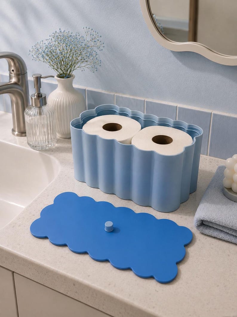 May include: A light blue rectangular toilet paper holder with a scalloped edge design, containing two rolls of white toilet paper. The holder includes a separate, matching dark blue lid with a small central knob for easy removal and placement.