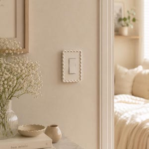 May include: A rectangular light switch cover plate featuring a scalloped, beaded edge design in a solid off-white finish. The cover plate is mounted on a wall and surrounds a standard vertical rocker-style light switch with a central rectangular toggle.