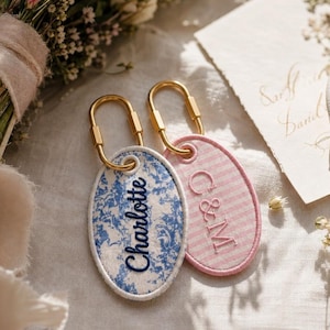 May include: Two oval-shaped fabric keychains with gold-toned metal carabiner clips. One keychain features a blue and white floral pattern with the name Charlotte embroidered in blue. The other has pink and white stripes with C&M embroidered in pink.