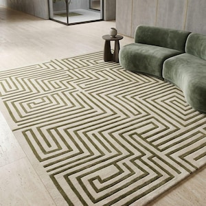 Könnte beinhalten: Ein rechteckiger Teppich mit einem erhabenen, olivgrünen geometrischen Labyrinth-Muster auf cremefarbenem Grund. Das Design besteht aus durchgehenden, miteinander verbundenen Linien, die ein labyrinthartiges Motiv über die gesamte Fläche bilden.