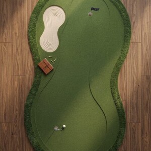May include: An oval-shaped indoor putting green rug with a deep green surface, a textured sand-coloured bunker, two holes with flags, a small white golf ball, and a brown leather-bound notebook with several golf tees resting on the side.