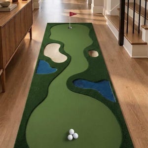 Peut inclure: Un tapis de putting d'intérieur rectangulaire avec une surface verte et trois balles de golf blanches au départ. Le tapis comprend deux obstacles d'eau bleus, un bunker de sable beige et un trou avec un drapeau rouge marqué de la lettre Y.