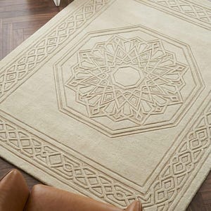 Könnte beinhalten: Ein rechteckiger, cremefarbener Teppich mit einem erhabenen, geschnitzten geometrischen Muster. Das Design umfasst ein zentrales achteckiges Medaillon mit komplexen sternartigen Motiven und eine breite Bordüre mit Gittermuster.