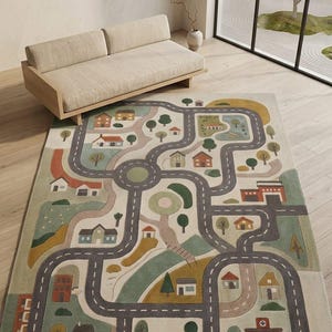 May include: A rectangular area rug featuring a printed town map design with winding gray roads, a central roundabout, and various illustrations of houses, trees, and buildings in muted tones of green, brown, orange, and blue on a cream background.