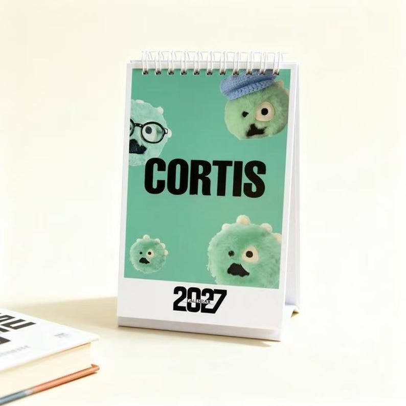 May include: A 2027 desk calendar with a spiral binding on a light green background. The cover features the text CORTIS in bold black letters and several fuzzy, light green, round creatures with eyes and mouths, one wearing a blue hat.