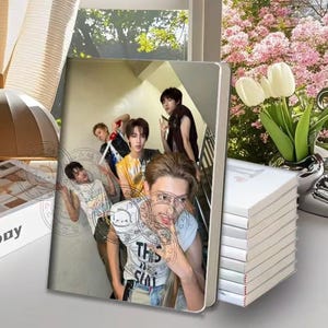 May include: A notebook featuring a photograph of four young people on the cover. The image shows them in a stairwell, with one person in the foreground wearing a white sleeveless shirt with text. A stack of white notebooks sits beside it.