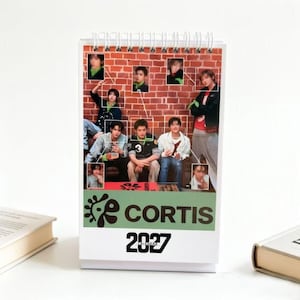 May include: A spiral-bound desk calendar for 2027 featuring a collage of photographs of a group of young people against a red brick wall background. The lower section displays the text CORTIS in bold black letters above the year 2027 in large font.